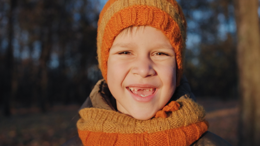 PORTRAIT: Happy Caucasian boy in an orange knitted hat and scarf. Child smiling with a mouth without front teeth. Concept of childhood and growing up. | Shutterstock HD Video #1084070413