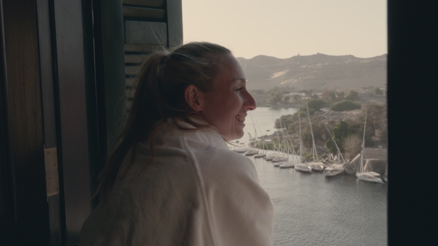 Slow motion: Young woman travelling wakes up in a hotel room and stands by the window enjoying the Nile in Aswan, Egypt 
