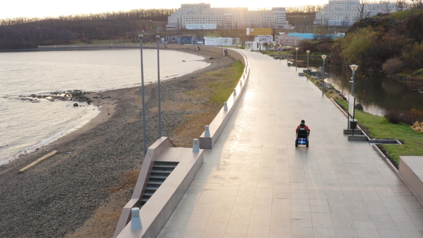 Aerial view of a man living with amputated legs and arms driving on modern electric wheelchair along the empty quay and sandy beach. Seaside. Autumn evening Royalty-Free Stock Footage #1084076152