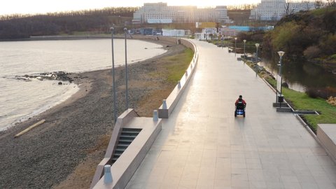 Aerial view of a man living with amputated legs and arms driving on modern electric wheelchair along the empty quay and sandy beach. Seaside. Autumn evening