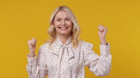 Excited jubilant happy elderly gray-haired blonde woman lady 40s years old wears pink shirt doing winner gesture celebrate clenching fists say yes isolated on plain yellow background studio portrait