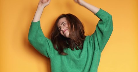 Emotions of people. Teen girl in green sweater dancing. Funny unusual brunette woman hair having fun, hands up, grimaces, smiling, dancing in studio against yellow background. Music, dance concept.