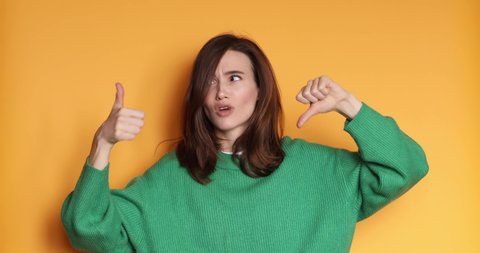 Young woman in green sweater making good-bad sign isolated on yellow background. Girl make choice, or makes a decision thumb up or tnumb down, like or dislike, yes or no. 