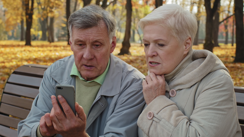 Upset elderly married couple sit on bench in autumn park read bad news on smartphone worried old people look at screen phone sad family feeling shocked read unpleasant message internet fraud concept Royalty-Free Stock Footage #1084085779