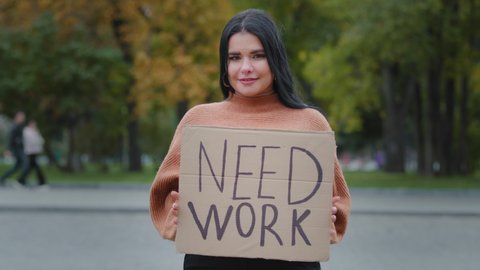 Close-up beautiful hispanic young woman stands on street near roadway unemployed girl holding cardboard banner NEED WORK female smiling shyly feeling awkwardness financial problems crisis dismissal