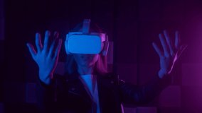 Young woman in futuristic VR headset gesturing with hand while interacting with cyberspace under neon light against black background