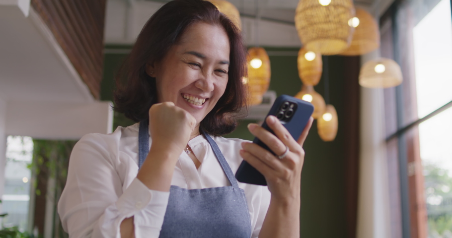 Asia mature adult woman people wear apron small cafe receive e-mail text good news SME lending service solution win seller store in social media app. Shock smile laugh face in sale order reopen shop. Royalty-Free Stock Footage #1084093819