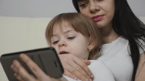 A dark-haired young mother and her little blonde daughter are sitting on the couch at home and using a smartphone to learn and communicate.