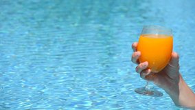 Female elegant hand holds glass goblet with cocktail or orange juice in blue pool. Summer pool party, concept of summer time vacation, fun, relaxation. Slow motion video. Copy space for your text.