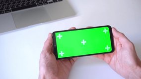 close-up. 4k. man holds smartphone horizontally with both hands, presses his finger on chroma key of green screen. on background of laptop in house. concept of people, technology, watching a movie