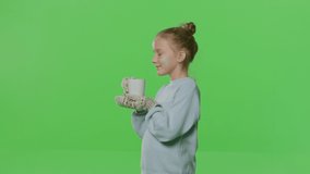 Pretty smiling girl drinking tea isolated on green screen background. Young white kid with hot drink smiling on Chroma key. 4k raw video footage slow motion