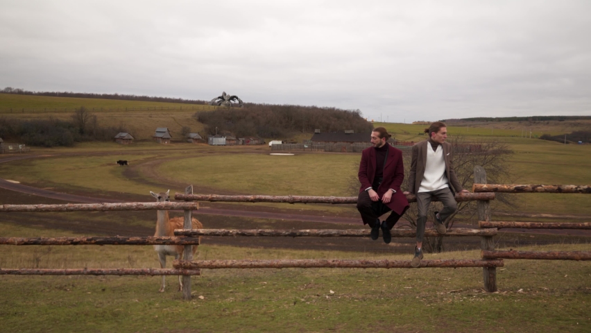 Siblings stand classic coat, sweater near wooden fence animal agricultural farm outside city. Cloudy autumn weather late autumn. Two businessmen, heirs business owners farm passed down from generation