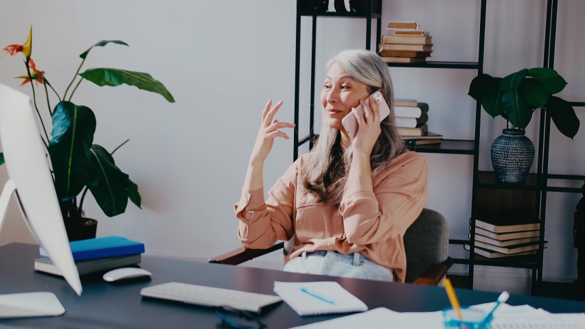 Attractive successful elderly businesswoman making phone call in modern office, sitting at desk with computer