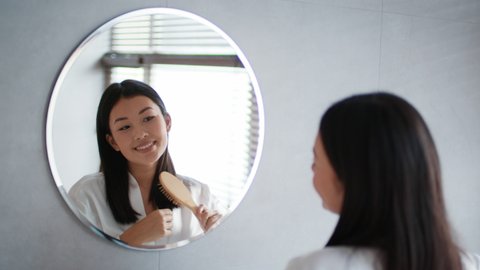 Happy Korean Woman Brushing Hair With Hairbrush Smiling Looking At Mirror Standing In Modern Bathroom At Home, Wearing White Bathrobe. Female Haircare And Cosmetics Concept. Selective Focus