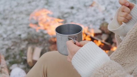 Woman holding roasted marshmallow and a metal cup of tea, enjoy the winter picnic with friends.