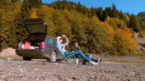A young guy and a girl are sitting on folding seats in a camping, drinking tea from metal mugs and looking at the mountains in autumn on a sunny day. There is a gas stove and a kettle nearby. car.