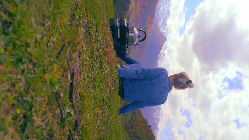 female traveler boils water in kettle on portable gas stove, there is mug next to it on grass, admires colorful view of autumn mountains, concept of camping and hiking in nature. vertical video,