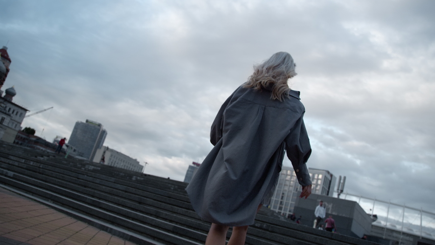 Young woman walking stairs at evening town in urban area. Back view female person running in big city landscape in cloud weather. Unknown girl in casual clothes going up stairs. City running concept.