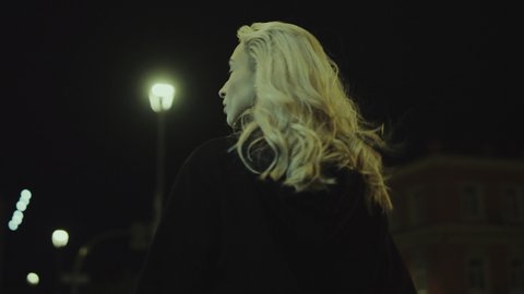 Blonde woman walking alone at late night in city lights. Attractive girl going to concert at night street. Back view confident female person wearing casual clothes while on a walk in slow motion. 