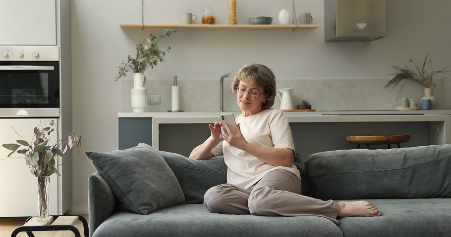 Positive middle aged lady on pension enjoy rest on sofa at home having fun with modern mobile phone test new application scroll social media content. Laughing mature female work online using cellphone Royalty-Free Stock Footage #1084126177