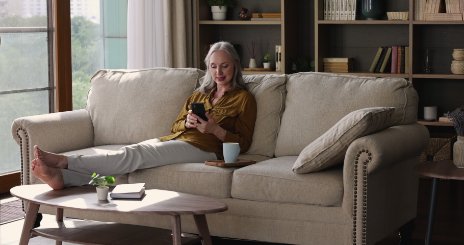 Beautiful senior adult female sit on sofa at cozy flat use mobile phone play game dial friend number to make video call. Old woman relax at home with smartphone networking online messaging in chat app Royalty-Free Stock Footage #1084126231