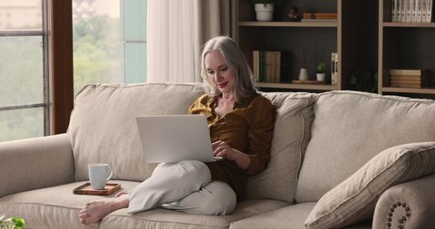 Relaxed woman retiree spend lazy weekend on sofa at home by laptop share news at social network contact with adult children in messenger app watch movie online. Elderly lady enjoy hot coffee using pc