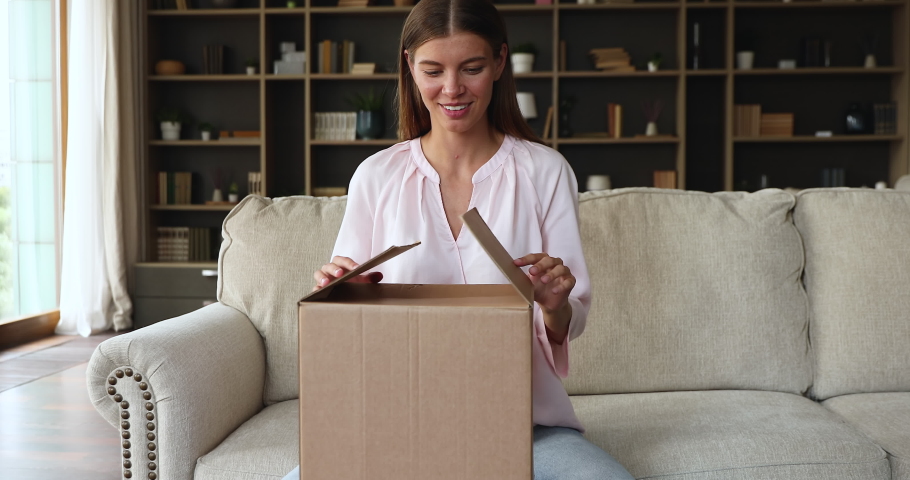 Smiling woman happy homeowner or renter, sit on sofa, opens received delivered parcel, unboxing carton box takes bought wicker baskets, storage jars, items for house interior decoration ordered online | Shutterstock HD Video #1084130455