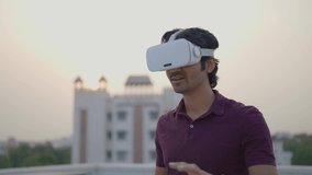 A young Indian modern male moving hands or hand gestures while having a first-time experience wearing a virtual reality headset or VR glasses standing outdoors on a rooftop of a building.