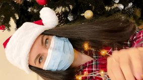 Vertical video. A girl in a Santa hat, in a medical mask and a garland around her neck records on video against the background of a Christmas tree. Christmas and new year concept in covid 19 setting.