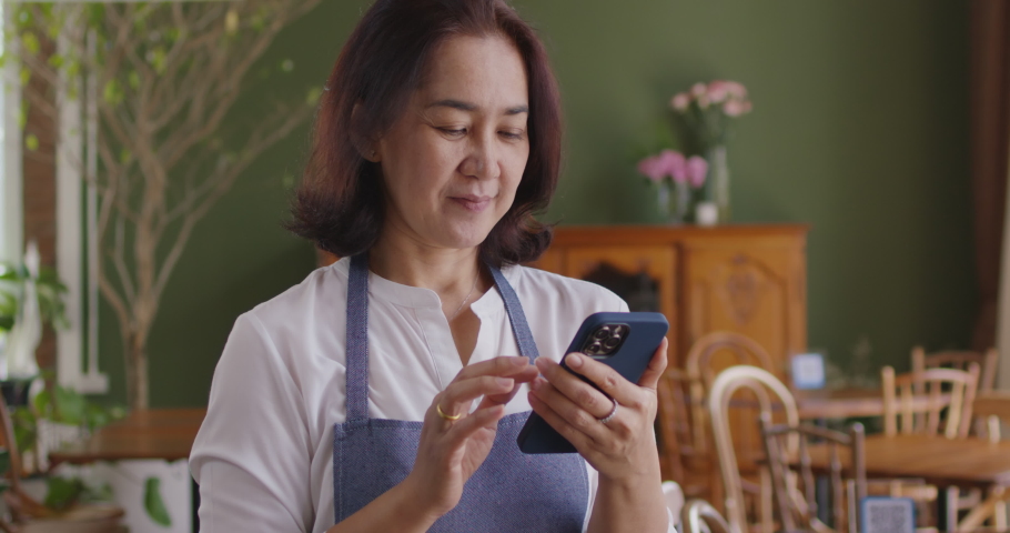 Asia mature adult woman people wear apron small cafe receive e-mail text good news SME lending service solution win seller store in social media app. Shock smile laugh face in sale order reopen shop. Royalty-Free Stock Footage #1084143001