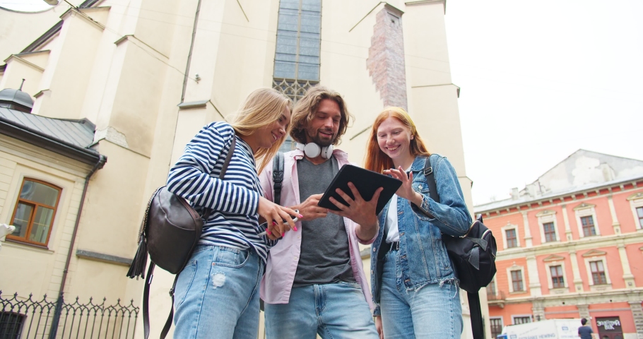 Young female and male students with bags standing on street and using gadget. Charming guy holding tablet and showing pictures to girl friends. Technology, friendship, education concept.