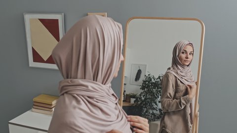 Beautiful Muslim woman putting on hijab and looking in mirror while getting dressed at home