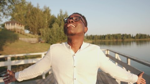 A young black man stands on the pier by the river and looks up at the sky with his arms outstretched. Freedom concept. Slow motion