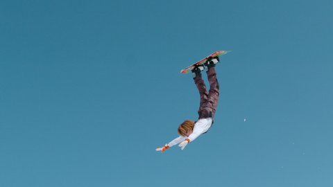 SLOW MOTION VERTICAL: Extreme snowboarder catches big air and does a spectacular backflip with outstretched arms. Action shot of a stoked male tourist doing a tumbling trick while snowboarding in Alps