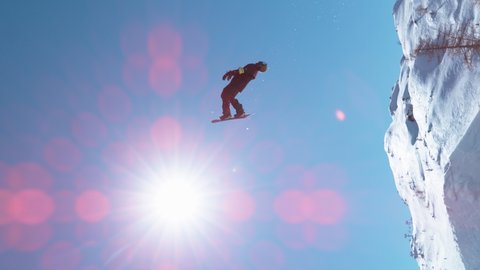 SLOW MOTION, VERTICAL, LENS FLARE: Bright winter sunbeams shine on a professional male snowboarder doing a backflip after taking off a kicker. Action shot of a freestyle snowboarder doing a flip.