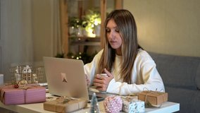 Happy and smiling young woman wrapping and preparing Christmas gifts, using laptop and having online video chat. Online shopping at Christmas holidays. Home interior with lights and plants