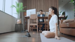 Working together at home,asian female wife workout yoga online training in living room while her caucasian male husband working using desktop at home in living room sit at background at home