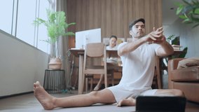 Working together at home,caucasian male husband work out yoga online training in living room while his asian female wife working using desktop at home in living room sit at background at home