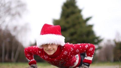 boy in a Christmas red sweater with a reindeer and a Santa hat in the park does push-up exercises