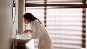Young Beautiful Asian Lady Wearing Silk Robe Washing Face In Bathroom, Attractive Korean Female Making Morning Hygiene At Home, Standing Near Sink And Splashing Water On Skin, Side View, Slow Motion