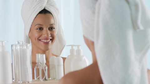 Gorgeous young slim African American woman with a towel on head and bare shoulders rubs hands and smells them enjoying the fragrance in front of the mirror in bathroom Hand care cosmetics commercial