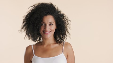 Beautiful African American young slim black-haired woman in white top turns to the camera touching her curly hair and smiling wide on beige background  Hair styling concept