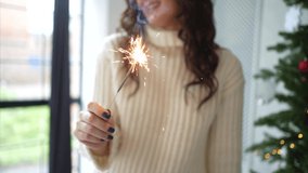 Video footage of Lovely young girl with long dark hair in sweater sits in armchair in cozy room with christmas tree and sparklers HD Slow motion.