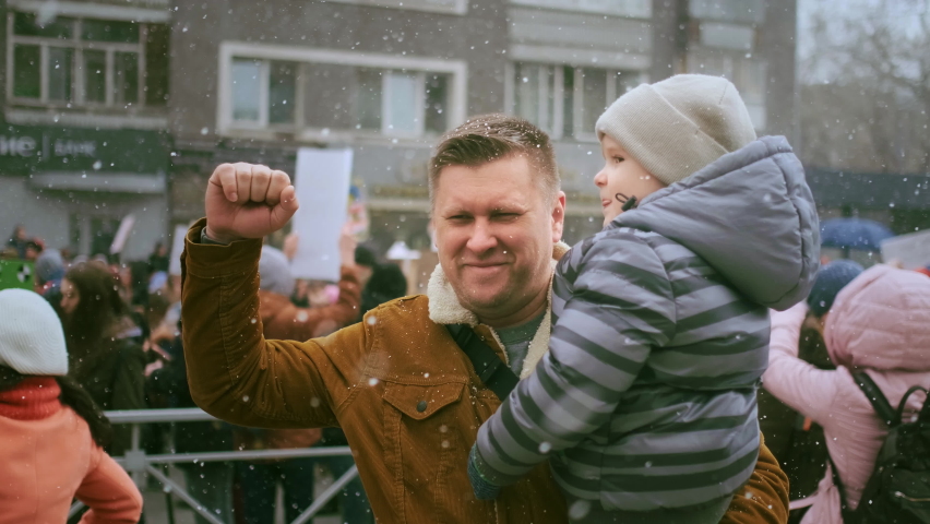 Single father protesting with his kid. Snowing weather during city opposition demonstration. Protester dad with child on arms. Man waves with raised fist. Political activists rally for dads rights. Royalty-Free Stock Footage #1084162780