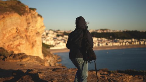 Tourist traveler woman with backpack stands on rocks of Nazare town. European tourism, traveling in Portugal. Mountains of Forte de Sao Miguel Arcanjo. Tourists take phone pictures, film sea or ocean.