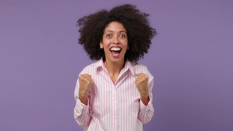 Excited jubilant overjoyed happy young black woman 20s years old wear pink shirt doing winner gesture celebrate clenching fists say yes isolated on plain pastel light purple background studio portrait