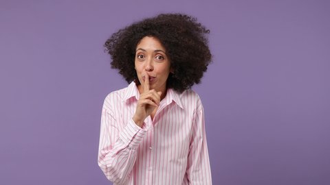Secret fashionable swanky young black woman 20s years old wear pink shirt look aside say hush be quiet with finger on lips shhh gesture isolated on plain pastel light purple background studio portrait