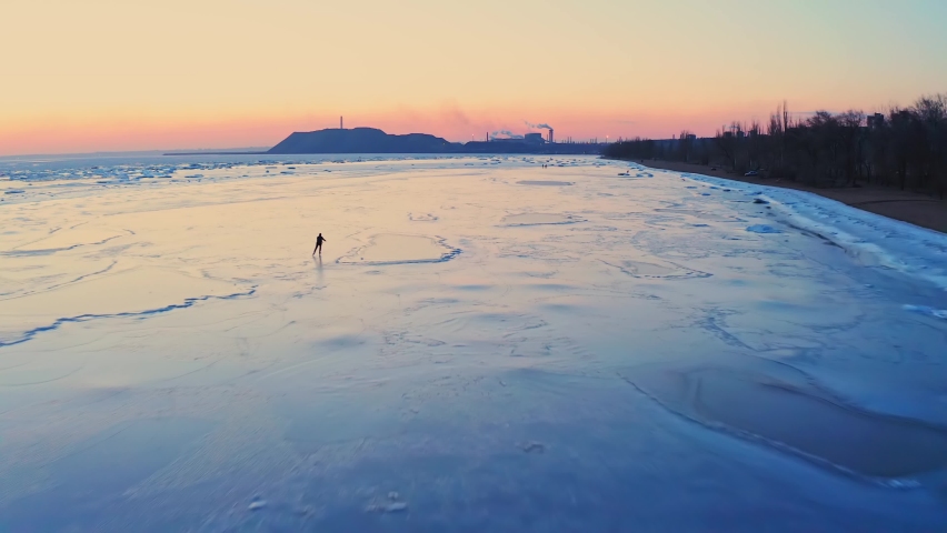 A man is skating on the ice of the frozen sea near the coast of the industrial town at sunset. Ice skating training. Winter leisure at the seaside. Drone footage.