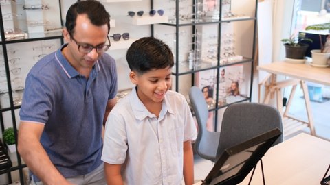Indian father hold glasses that his son wear and look to mirror with happiness during come to optical shop for buying new glasses.