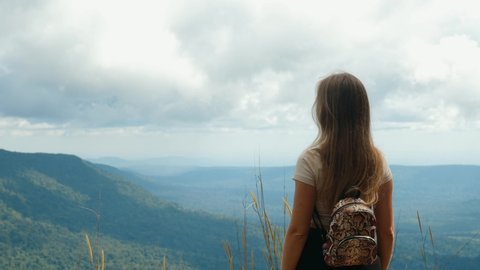 Inspiration travel. Back view of woman with backpack standing on top of mountain, enjoy summer nature and touch her long hair by hand. Happiness, wanderlust and freedom concept. 4k